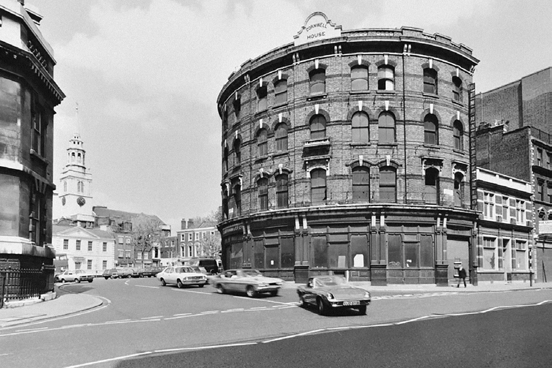 Archive photo of Cornwell House, a four story city building showing its round facade and corner position. Three cars in the foreground.